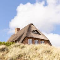 Ein traditionelles Reetdachhaus mit Ziegelmauerwerk und Gaube auf einer Düne, umgeben von Strandhafer unter blauem Himmel – eine begehrte Immobilienlage.
