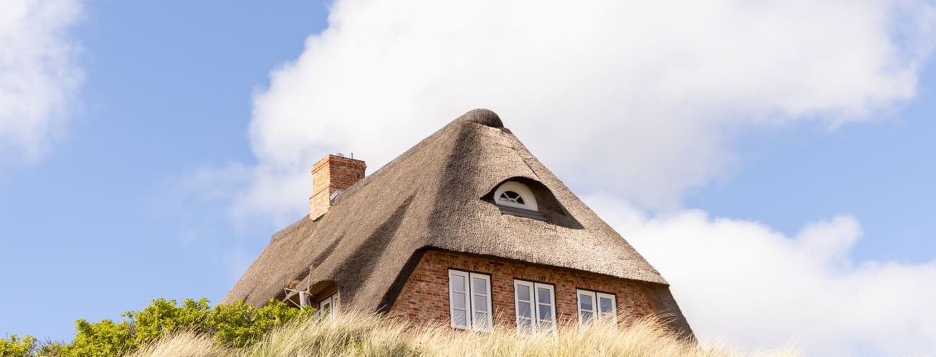 Ein traditionelles Reetdachhaus mit Ziegelmauerwerk und Gaube auf einer Düne, umgeben von Strandhafer unter blauem Himmel – eine begehrte Immobilienlage.