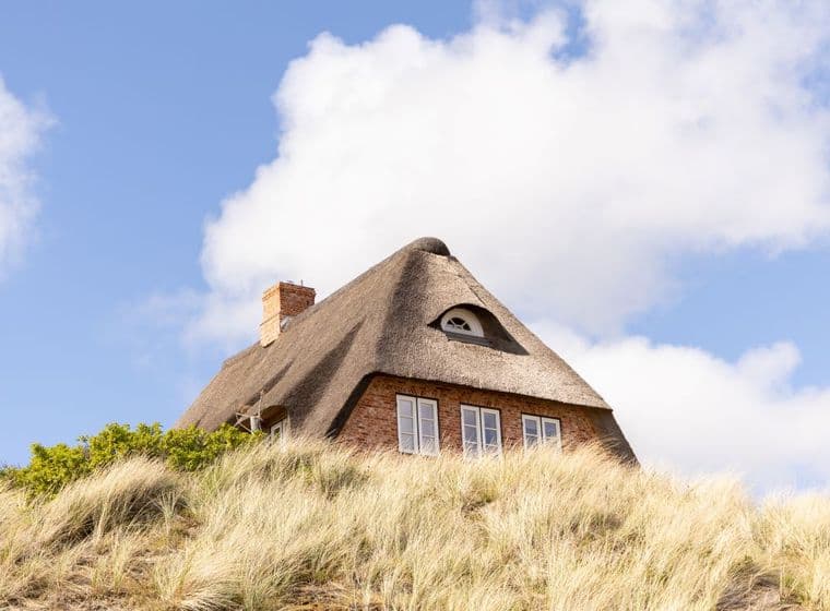 Ein traditionelles Reetdachhaus mit Ziegelmauerwerk und Gaube auf einer Düne, umgeben von Strandhafer unter blauem Himmel – eine begehrte Immobilienlage.