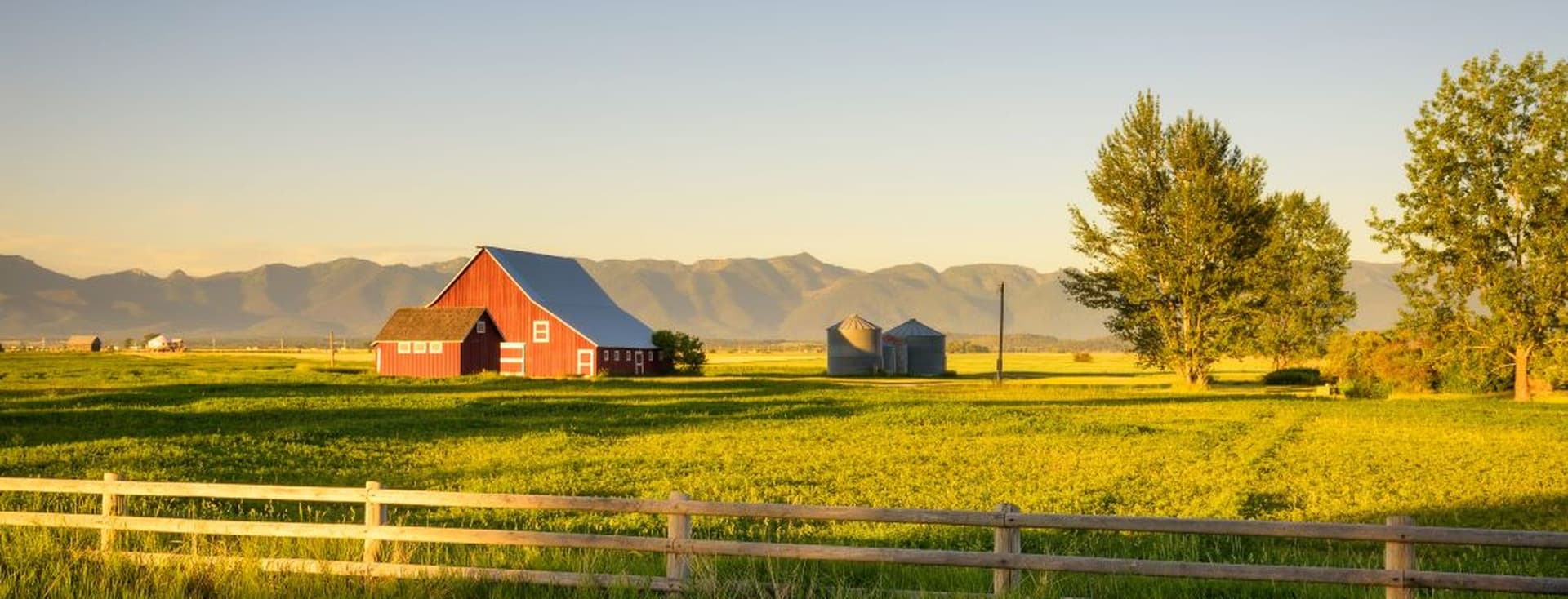 A red barn and silos sit in a green field under a clear blue sky with mountains in the background, bordered by a wooden fence.