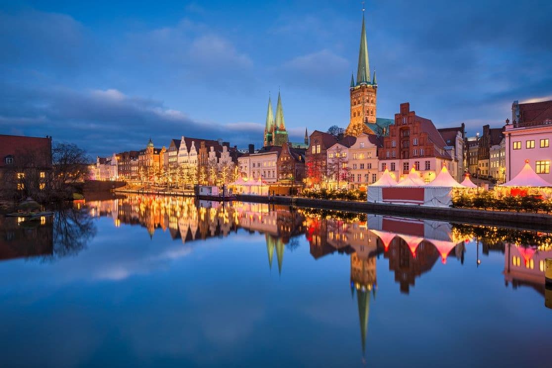 Scenic view of Lübeck in winter with illuminated buildings and church spires reflecting on a calm river at dusk.