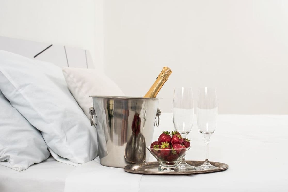 Tablett mit Sekt, Gläsern und Erdbeeren auf einem Hotelbett mit weißen Kissen