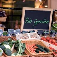 Ein Marktstand mit Biogemüse und einem Schild mit der Aufschrift „Bio Gemüse“. Die Körbe enthalten Tomaten, Gurken, Brokkoli und mehr.
