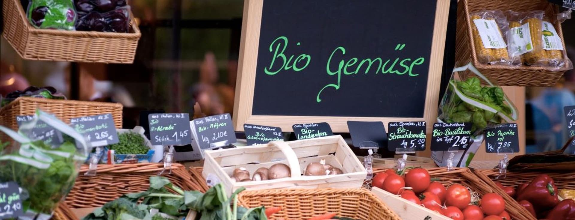 A market stall displaying organic vegetables with a sign reading "Bio Gemüse." Baskets contain tomatoes, cucumbers, broccoli, and more.