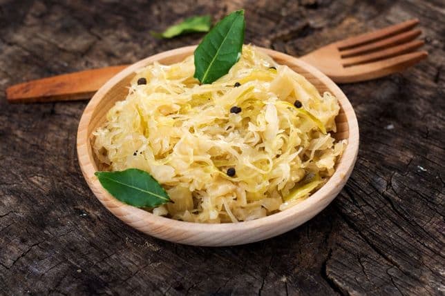 A wooden bowl of sauerkraut garnished with bay leaves and peppercorns, placed on a rustic wooden surface with a wooden fork beside it.