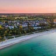 Luftaufnahme vom Timmendorfer Strand bei Sonnenuntergang mit einem von Sonnenschirmen gesäumten Sandstrand, grüner Landschaft und ruhigem türkisfarbenem Wasser.
