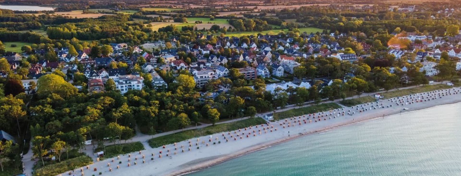 Aerial view of Timmendorfer Strand at sunset with a sandy beach lined with parasols, green landscape and calm turquoise water.