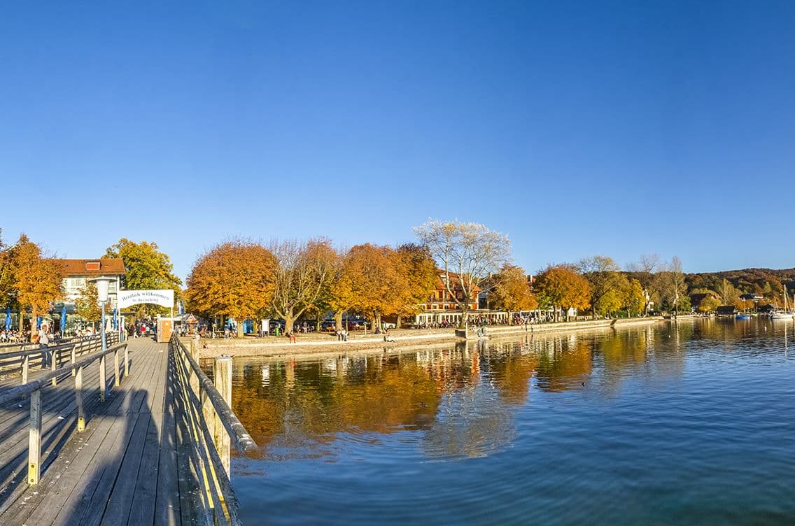 Wooden pier with railings leading to the busy lakefront in Herrsching at Lake Ammersee, with autumn trees and calm blue water