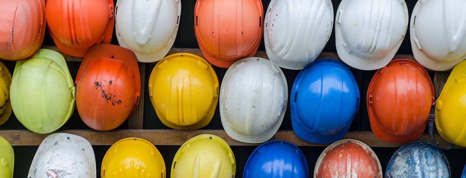 A wall of colorful hard hats, including white, yellow, orange, green, and blue, are neatly arranged on wooden shelves.