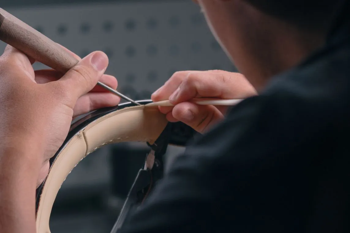 A close-up of hands sewing a curved piece of beige leather in a workshop using a needle and awl.