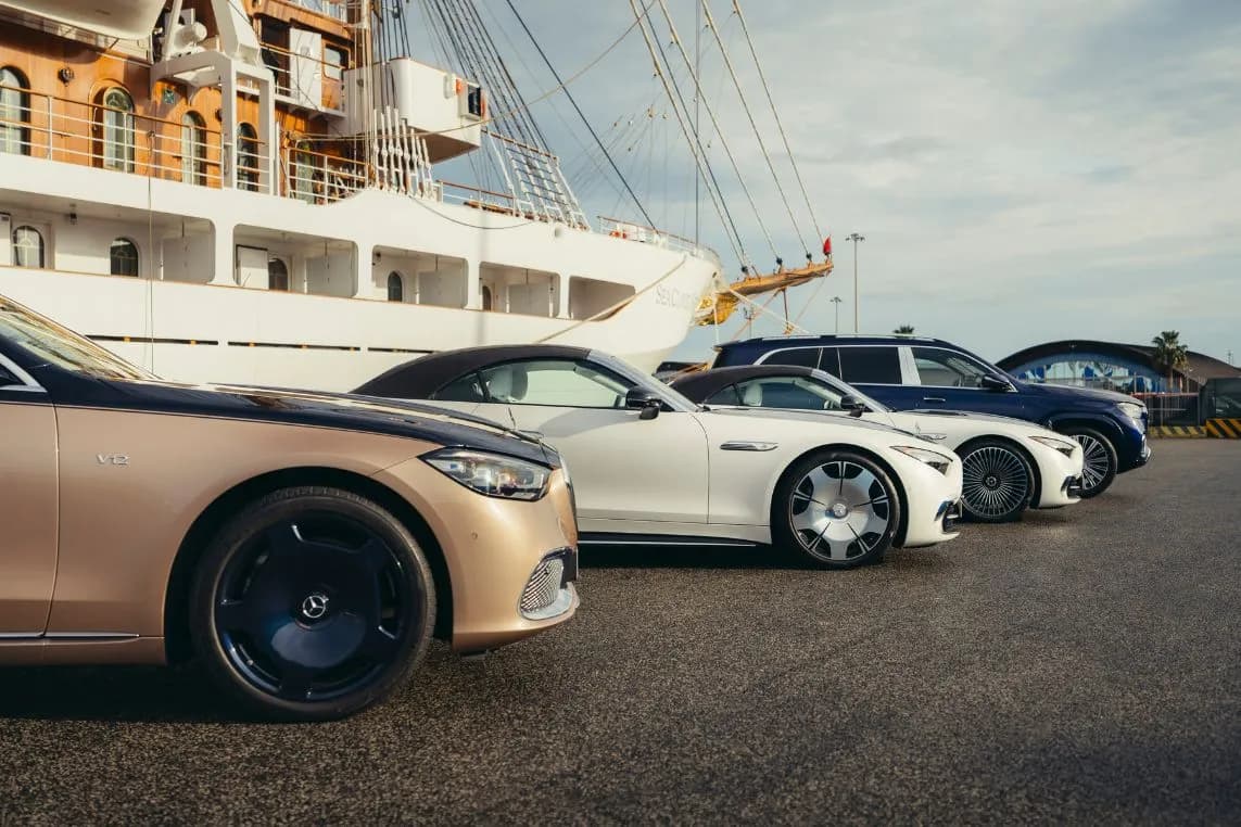 A row of luxury cars parked at a marina, behind which a large white ship with its rigging towers up.