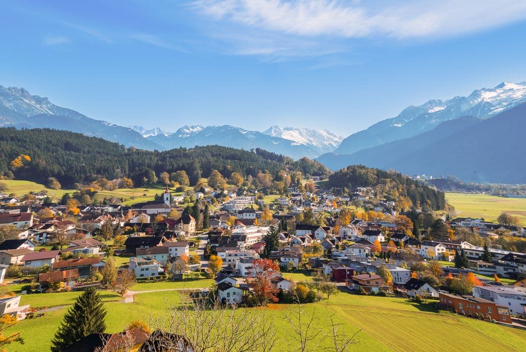 A picturesque village nestled in a valley with colorful houses, surrounded by lush greenery and snow-capped mountains under a clear blue sky.