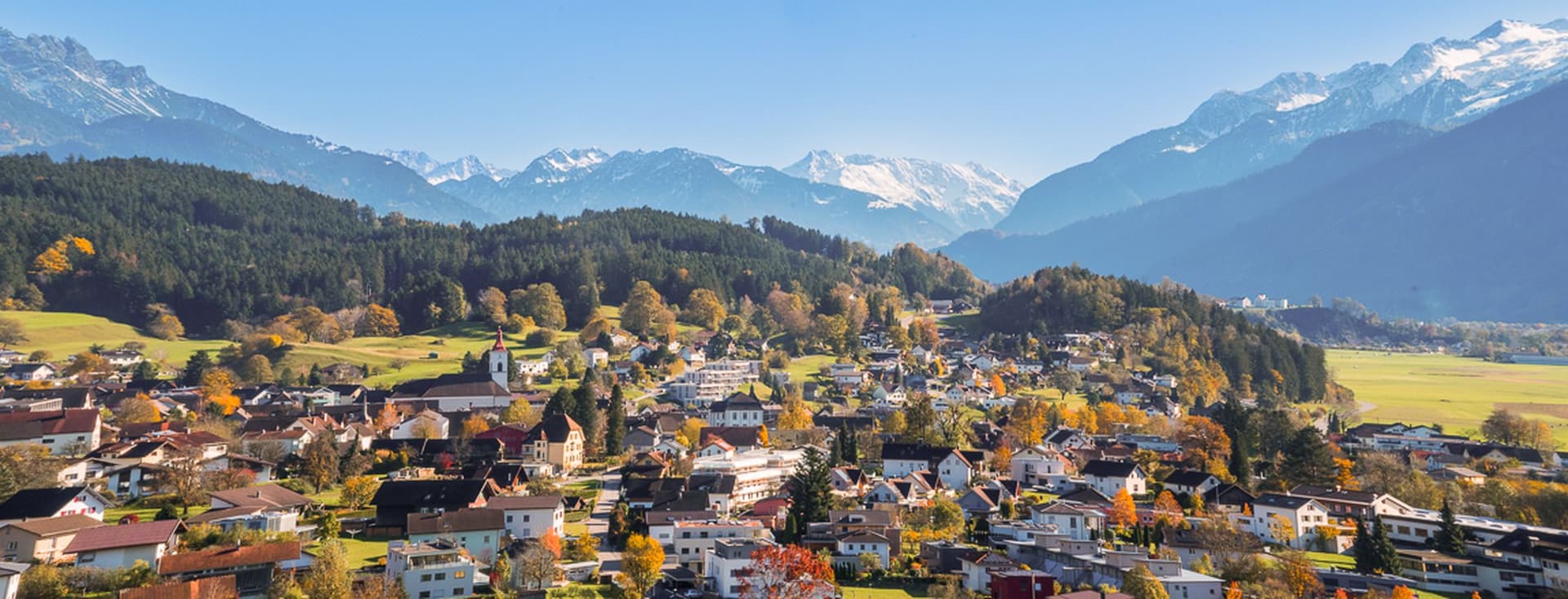 A picturesque village nestled in a valley with colorful houses, surrounded by lush greenery and snow-capped mountains under a clear blue sky.