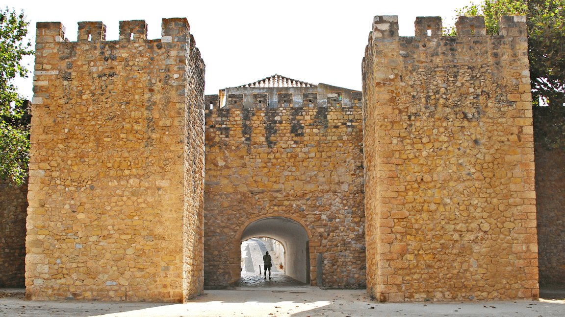 Ancient stone fortress entrance with two tall towers, arched passageway, and a person walking through. Sunlit and surrounded by trees.