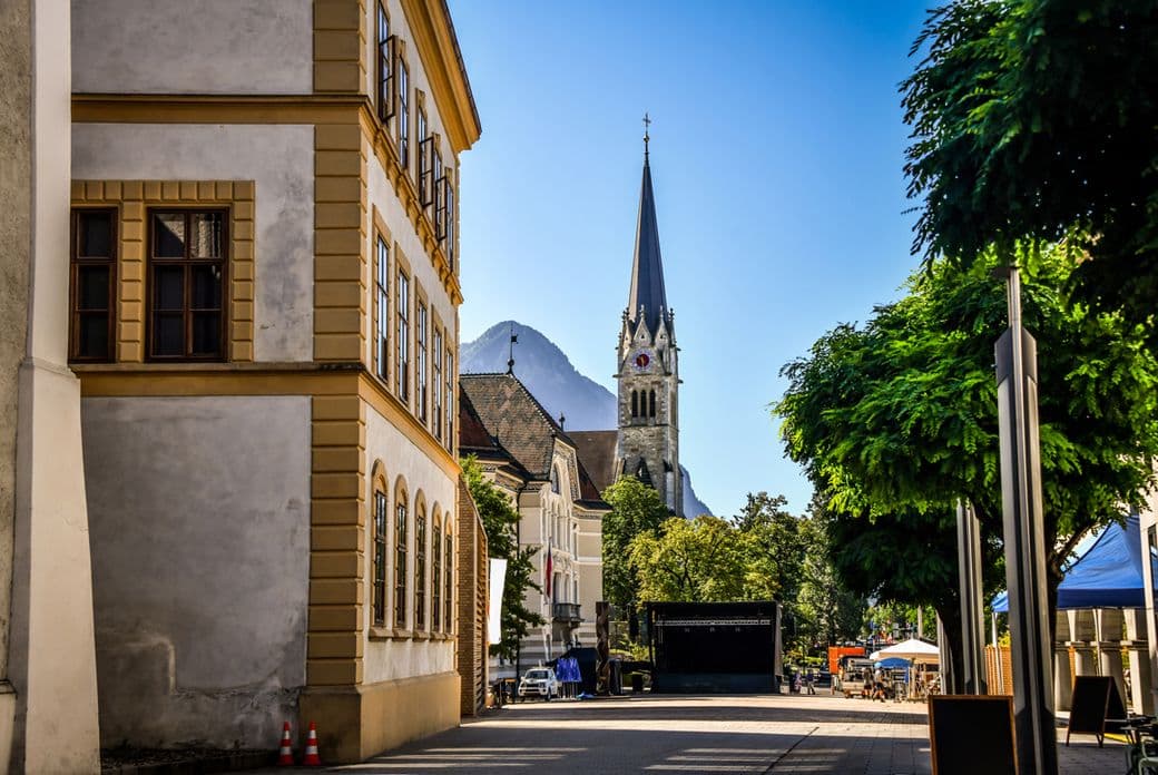 Street view of Vaduz, Liechtenstein, featuring a tall church spire, historic buildings, and a mountain backdrop under a clear blue sky.