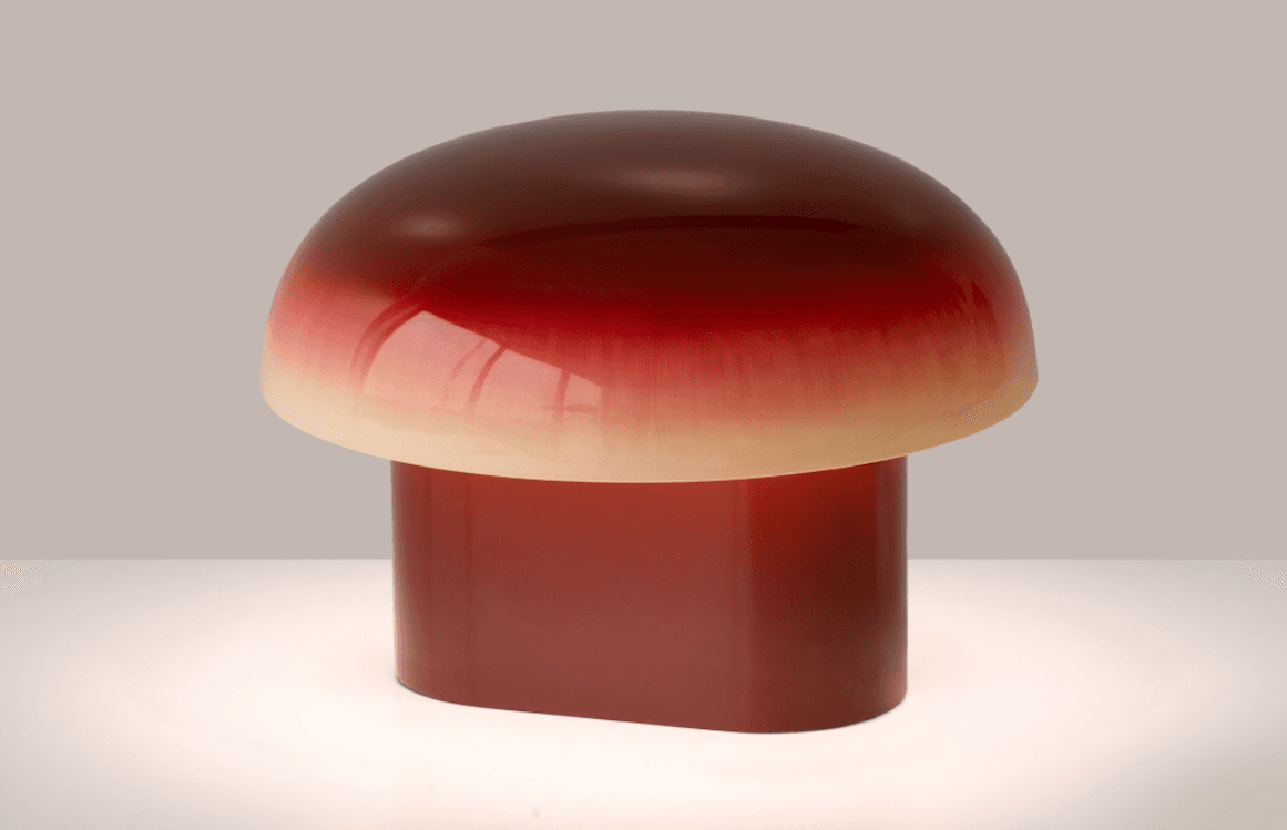 A mushroom-shaped lamp with a glossy gradient from deep red to cream, emitting a soft glow on a white surface.