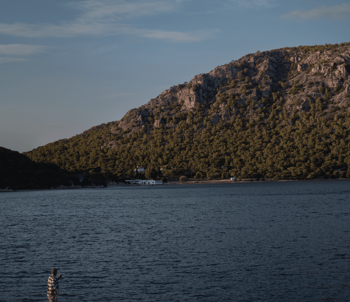 Scenic view of a mountain covered in green trees, overlooking a calm blue sea. A white building sits at the base of the mountain. A person stands in the foreground.