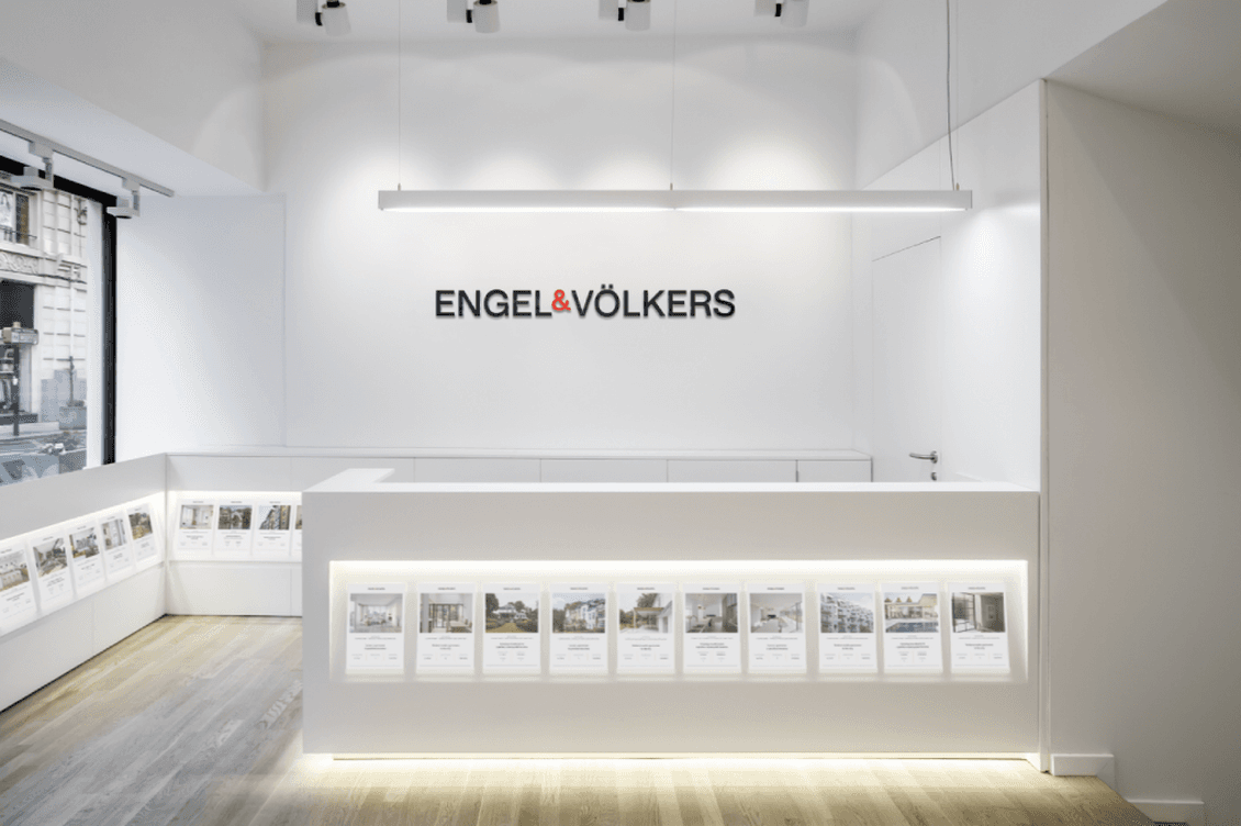Interior view of a minimalist Engel & Völkers real estate office featuring white walls, illuminated property displays along the walls, and the company logo mounted prominently on the back wall with modern lighting