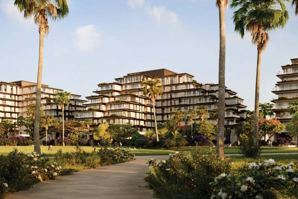 Pathway leading to modern, multi-story buildings surrounded by lush greenery and tall palm trees under a clear blue sky.