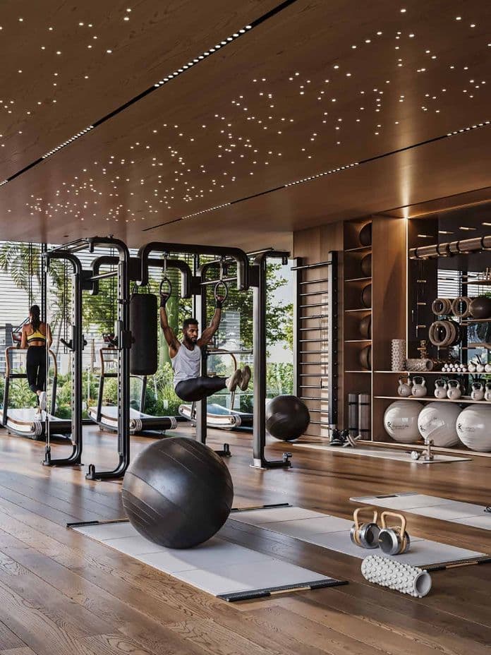 Modern gym with wooden flooring, exercise equipment, and a starlit ceiling. A person works out on rings, and another uses a treadmill.