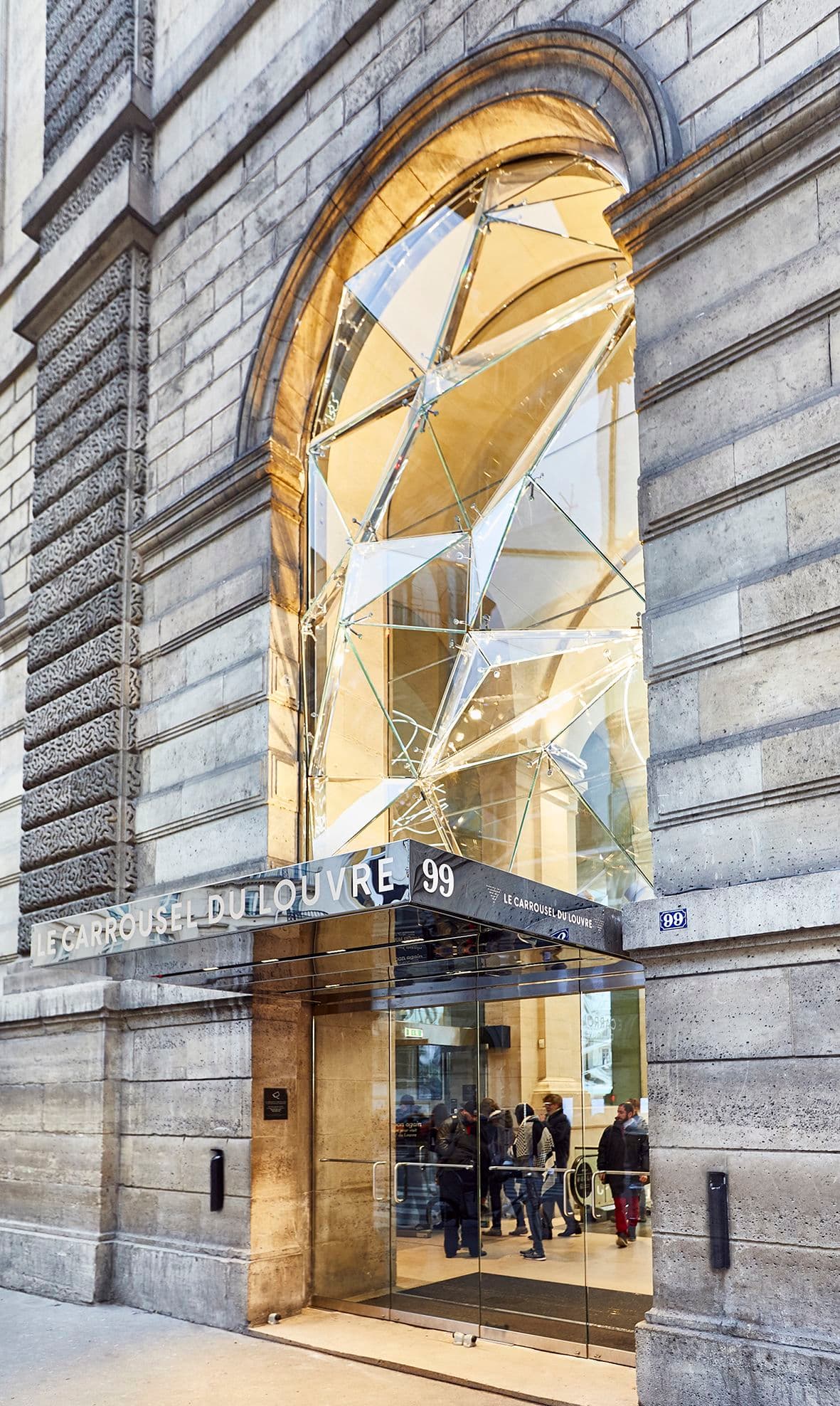 Vue extérieure de l'entrée du Carrousel du Louvre, avec une structure géométrique en verre au-dessus de la porte et des personnes qui entrent.