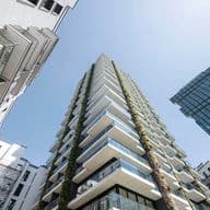 Low-angle view of a modern high-rise with staggered balconies, vertical greenery, and neighboring buildings against a clear blue sky.