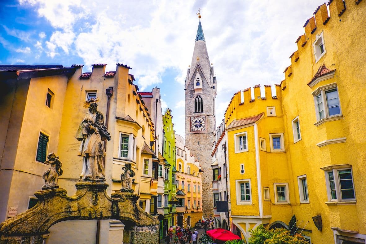 Farbenfroher europäischer Stadtplatz mit historischen Gebäuden, einem Glockenturm und einer Steinstatue. Menschen gehen unter einem blauen Himmel mit vereinzelten Wolken spazieren.