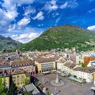Luftaufnahme eines europäischen Stadtplatzes mit farbenfrohen Gebäuden, einem zentralen Brunnen und üppig grünen Hügeln unter einem blauen Himmel mit Wolken.