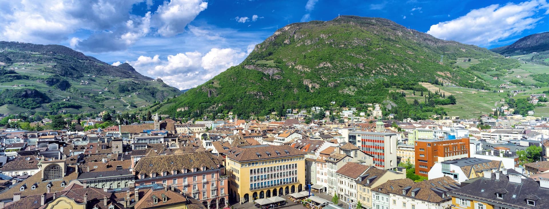 Luftaufnahme eines europäischen Stadtplatzes mit farbenfrohen Gebäuden, einem zentralen Brunnen und üppig grünen Hügeln unter einem blauen Himmel mit Wolken.