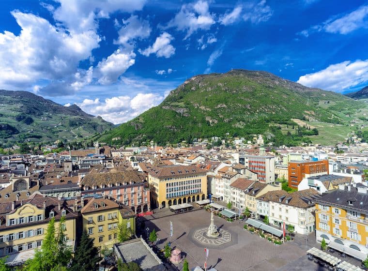 Luftaufnahme eines europäischen Stadtplatzes mit farbenfrohen Gebäuden, einem zentralen Brunnen und üppig grünen Hügeln unter einem blauen Himmel mit Wolken.