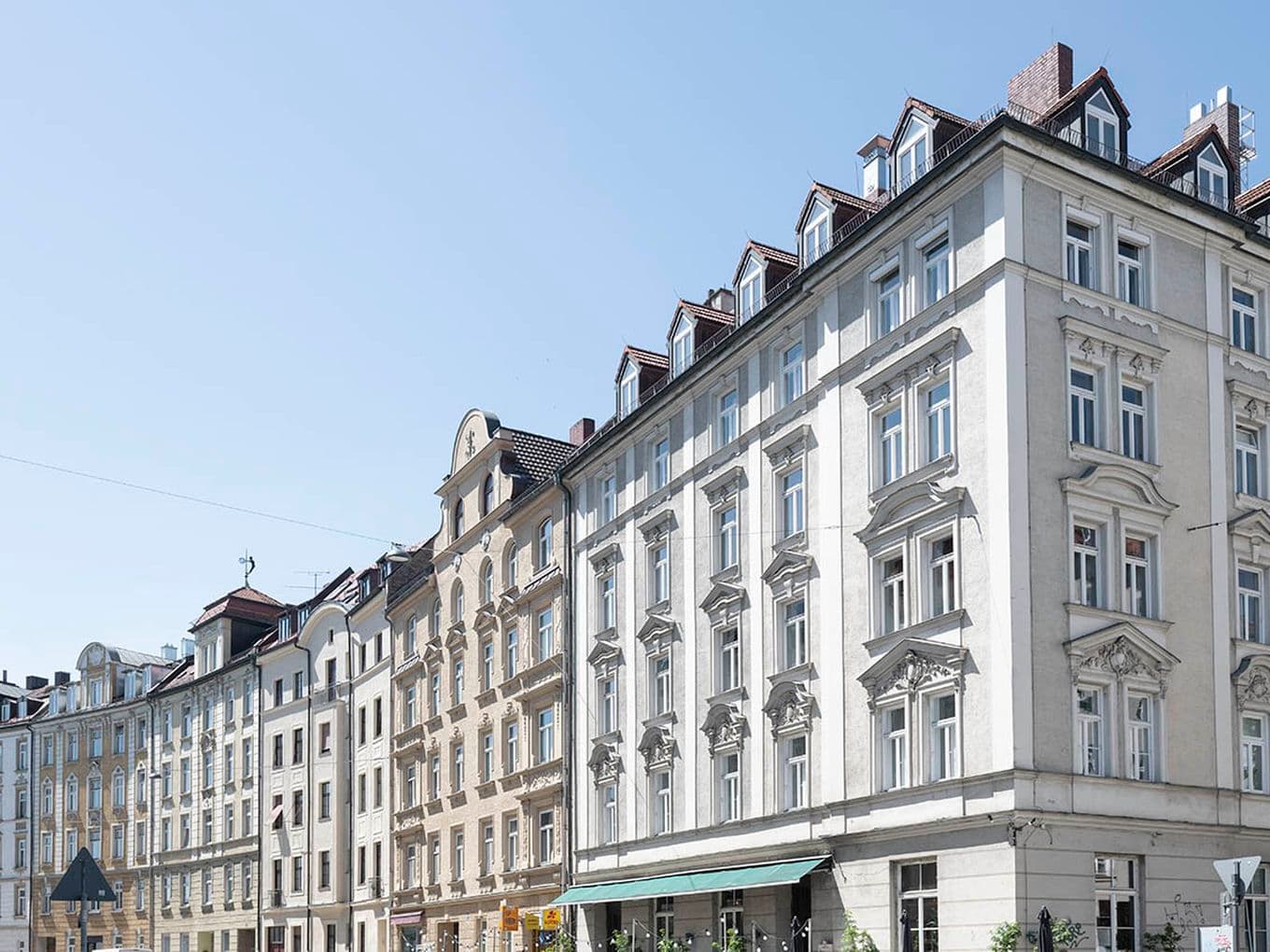 Row of elegant, historic European buildings with ornate facades under a clear blue sky. A café with outdoor seating is visible.