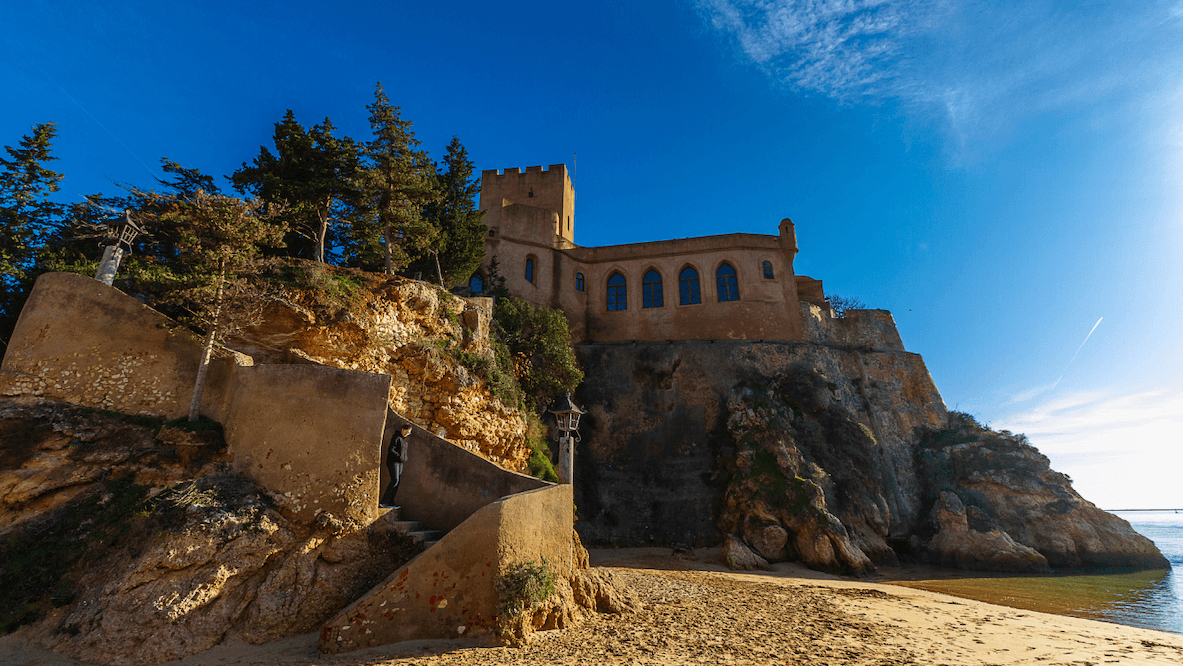 Historic castle on a rocky cliff by the sea, with a clear blue sky and sandy beach in the foreground.