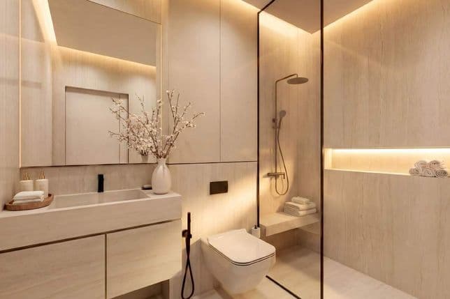 Modern bathroom with a glass shower, beige tiles, wall-mounted toilet, sink with a vase of branches, and towels on a shelf.