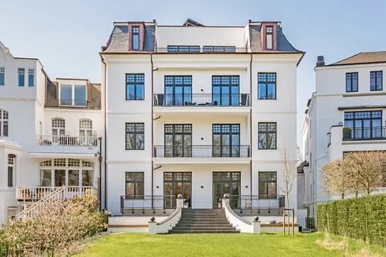 Weißes, mehrstöckiges Reihenhaus mit symmetrischen Fenstern und Balkonen sowie einer geschwungenen zentralen Treppe zum Eingang, umgeben von einem grünen Rasen.