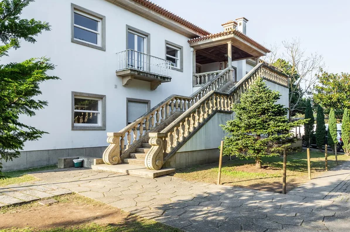 Ein zweistöckiges weißes Haus mit einer dekorativen Steintreppe, einem Balkon und einem kleinen Garten mit Bäumen. Sonniges Wetter und klarer Himmel.