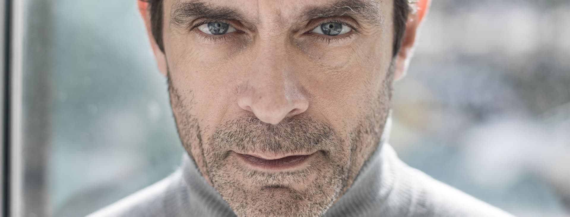 Φωτογραφία προσώπου ενός άνδρα με γαλανά μάτια και κοντά καστανά μαλλιά, που φοράει ένα γκρι ζιβάγκο πουλόβερ.