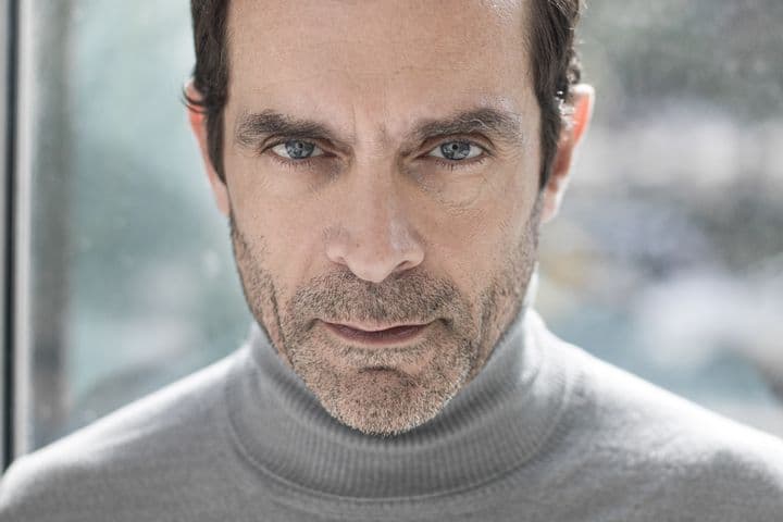 Φωτογραφία προσώπου ενός άνδρα με γαλανά μάτια και κοντά καστανά μαλλιά, που φοράει ένα γκρι ζιβάγκο πουλόβερ.