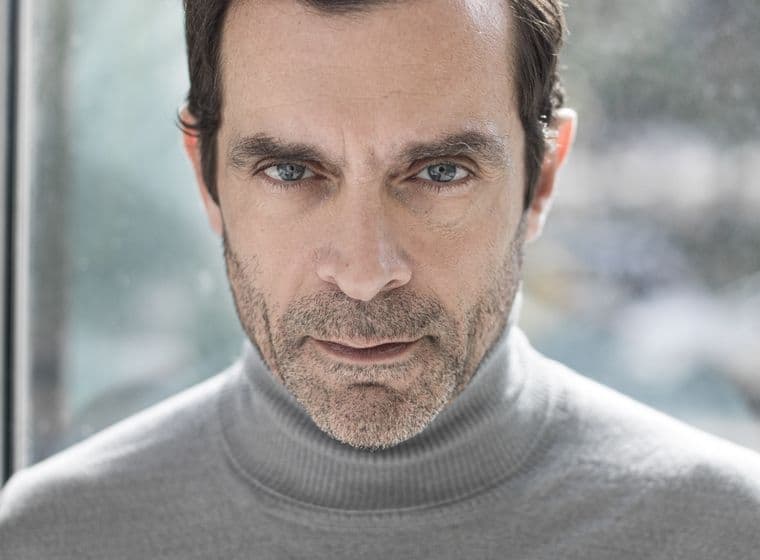 Φωτογραφία προσώπου ενός άνδρα με γαλανά μάτια και κοντά καστανά μαλλιά, που φοράει ένα γκρι ζιβάγκο πουλόβερ.