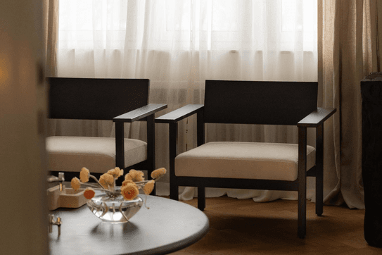 Cozy living room with two modern armchairs, a round table with flowers, and sheer curtains filtering soft light.