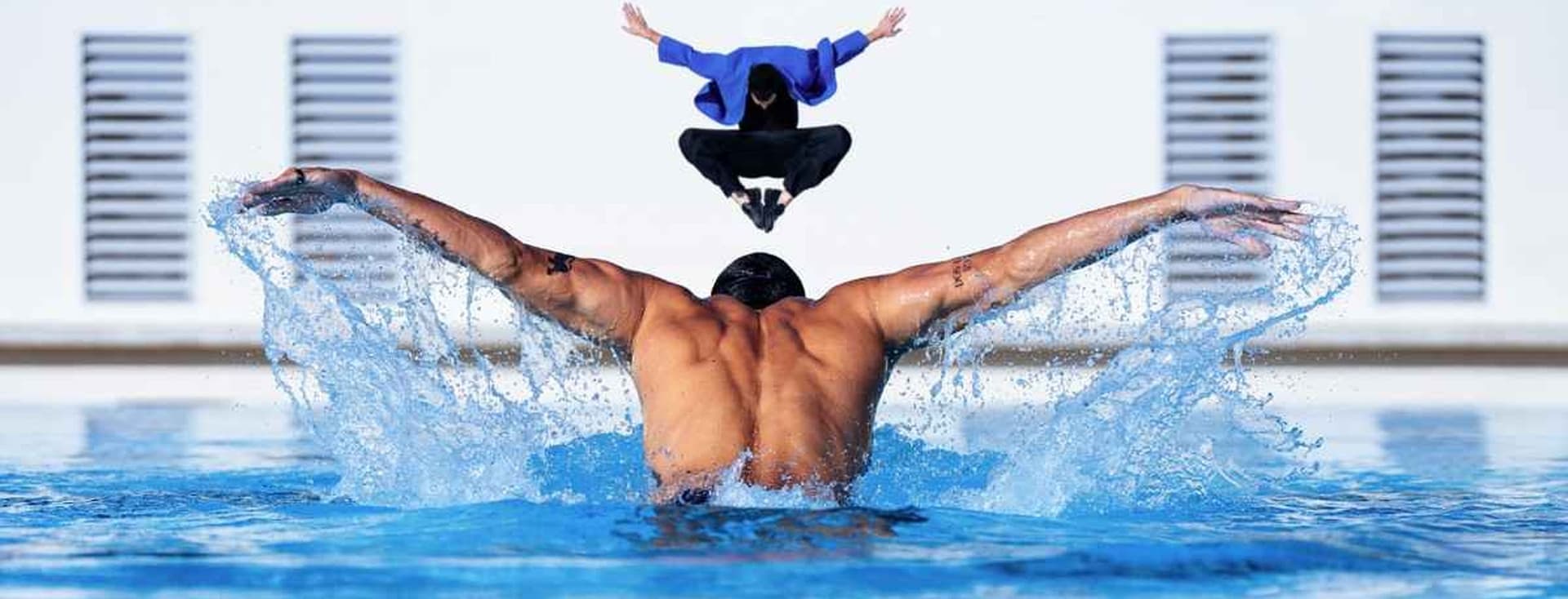 A muscular man swims butterfly in a blue pool, water splashing. Above him, a person in a blue jacket jumps. White building in background.