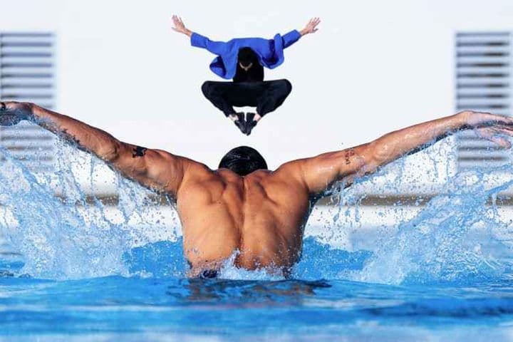 Un homme musclé nage le papillon dans une piscine bleue, l'eau éclaboussant. Au-dessus de lui, une personne en veste bleue saute. Bâtiment blanc en arrière-plan.