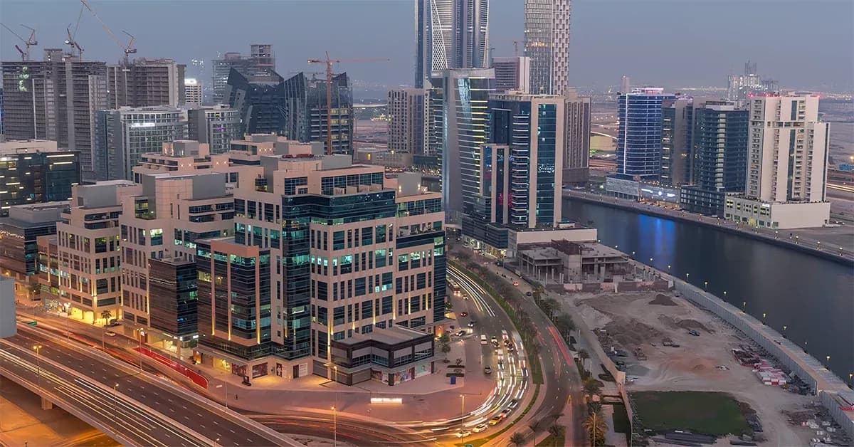 Dusk cityscape of modern skyscrapers and mid-rise office blocks along a curved waterfront canal, with streaking car lights on a busy road.