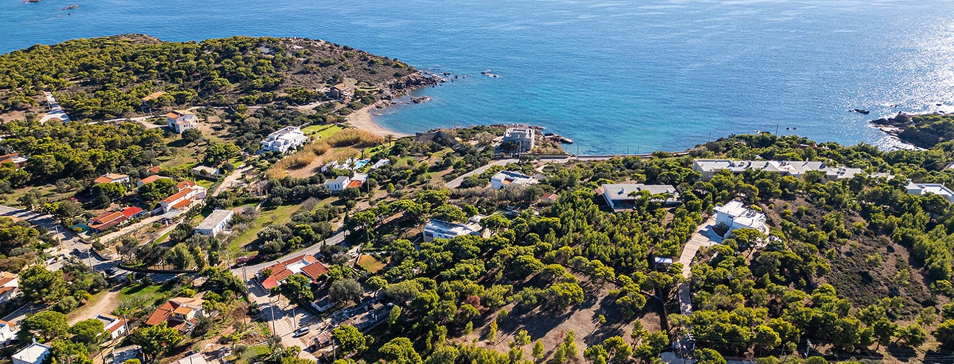 Αεροφωτογραφία μιας παραθαλάσσιας περιοχής με καταπράσινο τοπίο, διάσπαρτα σπίτια και καταγάλανα νερά. Ένας μικρός κόλπος είναι ορατός με μια βραχώδη ακτογραμμή. Ένα αθλητικό γήπεδο βρίσκεται στην κάτω δεξιά γωνία, περιτριγυρισμένο από δέντρα και σπίτια.