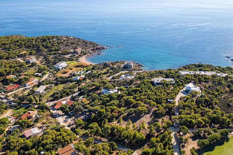 Αεροφωτογραφία μιας παραθαλάσσιας περιοχής με καταπράσινο τοπίο, διάσπαρτα σπίτια και καταγάλανα νερά. Ένας μικρός κόλπος είναι ορατός με μια βραχώδη ακτογραμμή. Ένα αθλητικό γήπεδο βρίσκεται στην κάτω δεξιά γωνία, περιτριγυρισμένο από δέντρα και σπίτια.