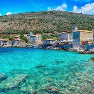 Coastal village with stone buildings by clear turquoise water, surrounded by rocky hills under a bright blue sky.