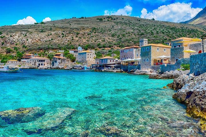 Coastal village with stone buildings by clear turquoise water, surrounded by rocky hills under a bright blue sky.