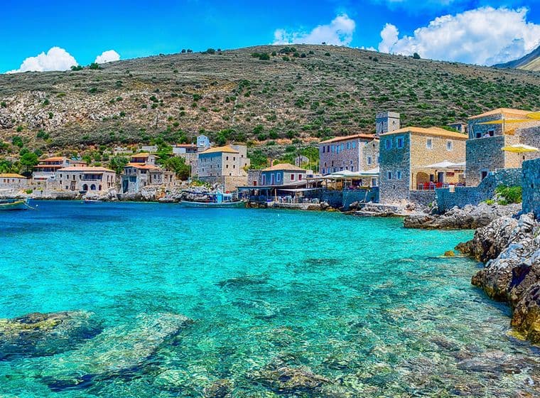 Coastal village with stone buildings by clear turquoise water, surrounded by rocky hills under a bright blue sky.