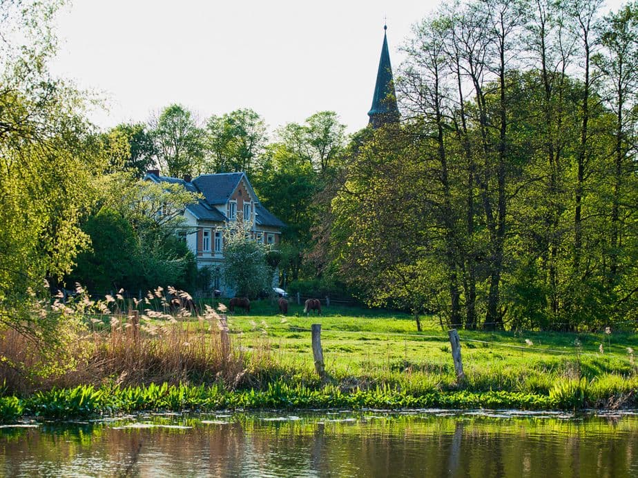 A tranquil landscape with a house and a church tower, surrounded by lush trees, next to a calm river in which the greenery is reflected.