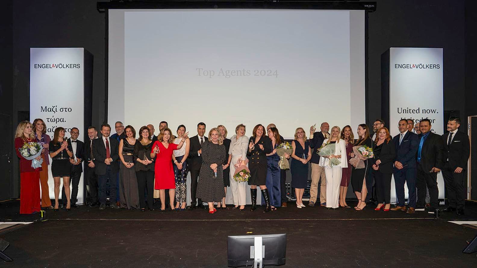 A group of people posing on stage for a photo, with a screen displaying "Top Agents 2024" behind them.