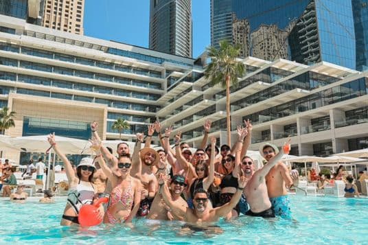 A group of people in swimwear cheerfully pose in a pool, surrounded by modern buildings and palm trees under a clear blue sky.